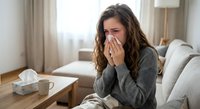 Young Caucasian woman with brown curly hair blowing nose into tissue while sitting on couch at home, feeling unwell with cold or flu symptoms in cozy living room.