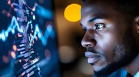 Young African American male trader analyzing financial data on computer screen with glowing charts and graphs in modern trading office environment.