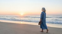 Senior woman in denim coat walking on sandy beach at sunrise, holding bag, ocean waves in background, peaceful coastal morning scene.