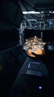 Hand holding glowing geometric network wireframe in dark tech laboratory environment with computer equipment and cables overhead, representing digital innovation and data connectivity.