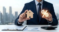Professional businessman in dark suit holding wooden house model and glowing percentage symbol representing mortgage rates and real estate investment opportunities.