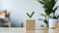 Green checkmark symbol floating above wooden cube on desk with blurred plant decor background, representing approval, success, or task completion.