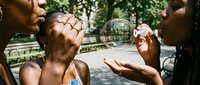 Young diverse friends enjoying outdoor summer day blowing soap bubbles in urban park setting with joyful expressions and casual lifestyle moments.