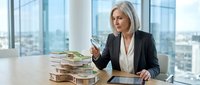 Middle-aged woman with white hair, Caucasian ethnicity, wearing professional business attire, sitting at a modern office desk with stacked currency and a tablet, analyzing financial data in a high-rise corporate setting.