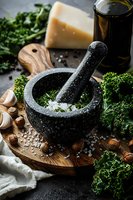 Black granite mortar and pestle with sea salt on wooden board, surrounded by fresh kale, parmesan cheese, garlic, olive oil and hazelnuts in moody lighting.