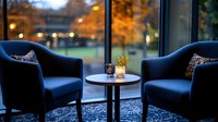 Cozy lounge area with two navy blue armchairs and round table by large window overlooking autumn park at dusk, soft ambient lighting creates intimate atmosphere.