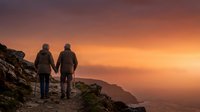 Two hikers holding hands stand on a mountain ridge at sunset, gazing at golden-orange sky and misty valleys below, capturing adventure and connection in nature.