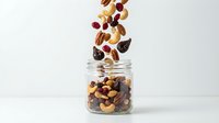 Mixed nuts and dried fruits falling into glass jar on white background, isolated for healthy snack packaging and nutrition advertising.