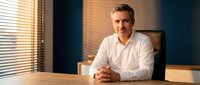 Professional middle-aged Caucasian businessman in white shirt sitting at office desk with warm lighting from window blinds for corporate presentations.