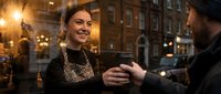 Young woman with dark hair and fair complexion, wearing a dark lace top, smiling while holding hands with a man during evening in an urban setting with warm street lighting and historic architecture.