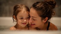 Young Caucasian mother and daughter enjoying intimate bonding moment together in warm bubble bath, sharing tender embrace and gentle smiles in cozy bathroom setting.
