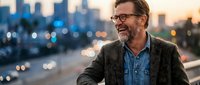 Mature businessman with glasses smiling outdoors at sunset with city skyline and bokeh lights in background. Professional corporate lifestyle concept.