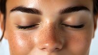 Close up portrait of young woman with freckles, natural makeup and closed eyes showing glowing healthy skin and long eyelashes against neutral background.