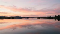 Serene lake at sunrise with pink and orange sky reflecting on calm water surface, distant mountains and tree silhouettes.