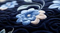 Intricate blue and white floral embroidery on dark fabric with detailed stitching patterns and decorative swirls for textile and craft projects.