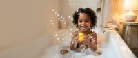 Happy African American toddler girl playing with rubber duck and soap bubbles during bath time in cozy bathroom with warm lighting for family content.