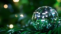 Shiny disco ball nestled among green palm leaves with soft bokeh lights in background creates nostalgic retro party atmosphere.