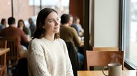 Young woman in beige sweater sitting at cafe table by window, smiling thoughtfully. Cozy coffee shop atmosphere with blurred people in background.