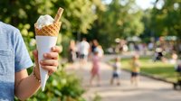 Cropped view of person holding vanilla soft serve ice cream cone outdoors in sunny park setting with blurred crowd and green trees in background, suitable for summer, food, and lifestyle content.