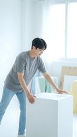 Young Asian man in casual gray shirt and jeans moving white furniture box in bright modern room with natural window light for relocation concept.