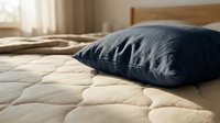 Dark blue pillow resting on quilted beige bedding in a bright bedroom with natural light, suitable for home decor and bedding product marketing.