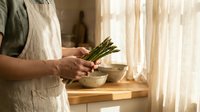 Woman in linen apron holding fresh asparagus spears in bright kitchen with wooden counter and soft natural lighting for healthy cooking concepts.