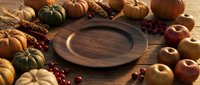 Autumn harvest arrangement with empty wooden plate surrounded by pumpkins, gourds, apples and cranberries on rustic wood surface for fall seasonal dining and Thanksgiving table styling.