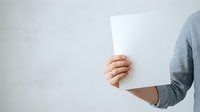 Person holding blank white paper sheet against neutral background, cropped torso view wearing casual gray long-sleeve shirt, suitable for mockup and advertising designs.