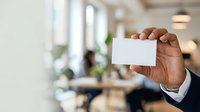 Young professional man of African descent displaying blank business card in modern office environment with colleagues working in background.