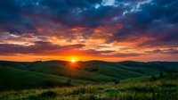 Dramatic sunset over rolling green hills with golden sun on horizon, vibrant orange and purple clouds illuminating rural landscape for nature and travel themes.