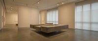 Modern minimalist gallery interior with concrete display platform and natural lighting from large windows creating serene exhibition space.