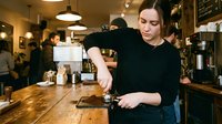 Young Caucasian female barista preparing coffee at espresso machine in busy cafe with warm lighting and customers in background.