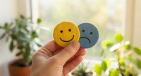 Hand holding yellow happy and gray sad emoji faces representing contrasting emotions and mental health awareness in bright indoor setting.