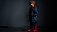 Young boy with curly red hair wearing navy raincoat and bright red rubber boots standing against dark background, ready for rainy weather adventure.