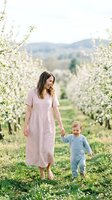 Young mother in pink dress walking with toddler son through blooming apple orchard in spring countryside for family lifestyle content.