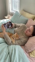 Young woman relaxing in bed with orange tabby cat while video calling on smartphone in cozy bedroom with pastel pillows and natural window light.