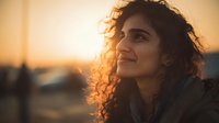 Young Middle Eastern woman with curly hair smiling at golden hour sunset, warm lighting creating peaceful moment outdoors for lifestyle content.