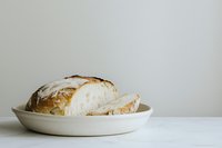Fresh crusty artisan sourdough bread loaf sliced on white ceramic plate against neutral background, minimalist food photography composition.