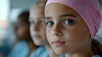 Young girl wearing pink headscarf with bright blue eyes looking forward with hope and determination in medical or healthcare setting.