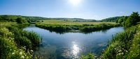 Scenic river landscape with lush green vegetation, rolling hills, and clear blue sky reflecting in calm water, suitable for nature and travel content.