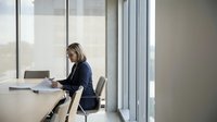 Professional businesswoman working on laptop in modern office conference room with large windows and natural light for corporate presentations.