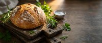 Freshly baked artisan bread loaf on rustic wooden cutting board with herbs and salt, perfect for bakery marketing and culinary content creation.