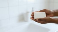 Person with dark skin tone holding white bar of soap in bathroom setting, promoting hygiene and personal care products.