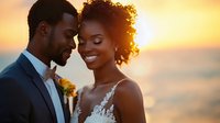 Young African American couple in wedding attire share intimate moment at sunset, with bride in lace dress and groom in tuxedo against golden sky background.
