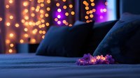 Cozy bedroom with soft fairy lights glowing on blue bedding, creating romantic atmosphere. Purple and orange bokeh lights in background add dreamy ambiance.