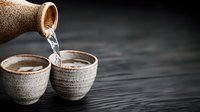 Pouring sake from traditional ceramic bottle into rustic cups on dark wooden table, dramatic lighting creates moody atmosphere for Japanese dining experience.