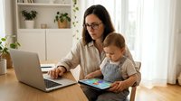 Working mother with toddler using laptop at home office desk while child reads colorful book, multitasking parent balancing career and childcare responsibilities.