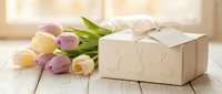 Elegant gift box with ribbon and fresh tulips on white wooden table, spring celebration concept for Mother's Day, Easter, birthday or anniversary occasions.