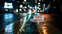Fake news paper sign on wet asphalt street with blurred city lights and traffic bokeh background at night creating urban misinformation concept.