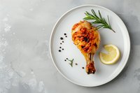 Grilled chicken leg with crispy golden skin served on white plate with fresh rosemary, lemon slice and black peppercorns on gray concrete background.